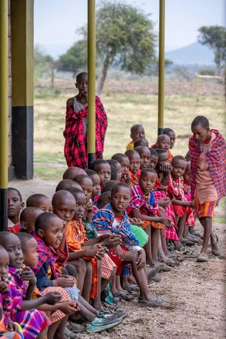 Maasai schoolchildren in bright shúkà seated in a long row along the veranda of a community school, with rangeland and hills behind.